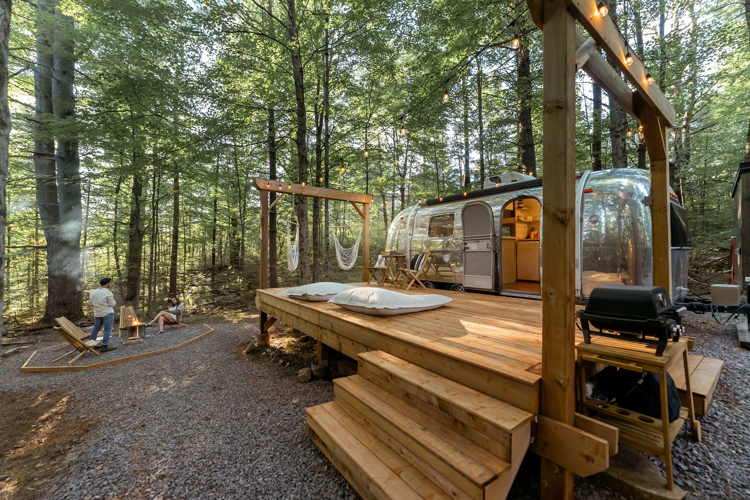 Un camping arbolado con una terraza de madera, una parrilla y una caravana.