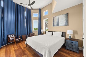 A bedroom with a large bed, a ceiling fan, a nightstand with a lamp, a chair, and a window with curtains. - Fully Renovated 5 BDR PCB House-Walk to Beach! (Panama City Beach)