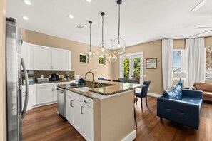 A modern kitchen with white cabinets, granite countertops, and a central island. There is a dining area with a blue sofa and a coffee table. - Fully Renovated 5 BDR PCB House-Walk to Beach! (Panama City Beach)