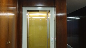 An elevator with a metallic interior and a glass door, surrounded by wooden walls and a tiled floor.