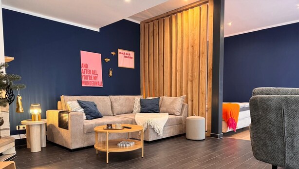 Una sala de estar moderna con una pared de acento azul oscuro, un separador de madera, un sofá color beige, una mesa de centro redonda y un sillón gris.