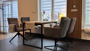 Una sala de estar moderna con piso de madera oscura, una mesa de comedor con sillas y un ventanal con persianas.