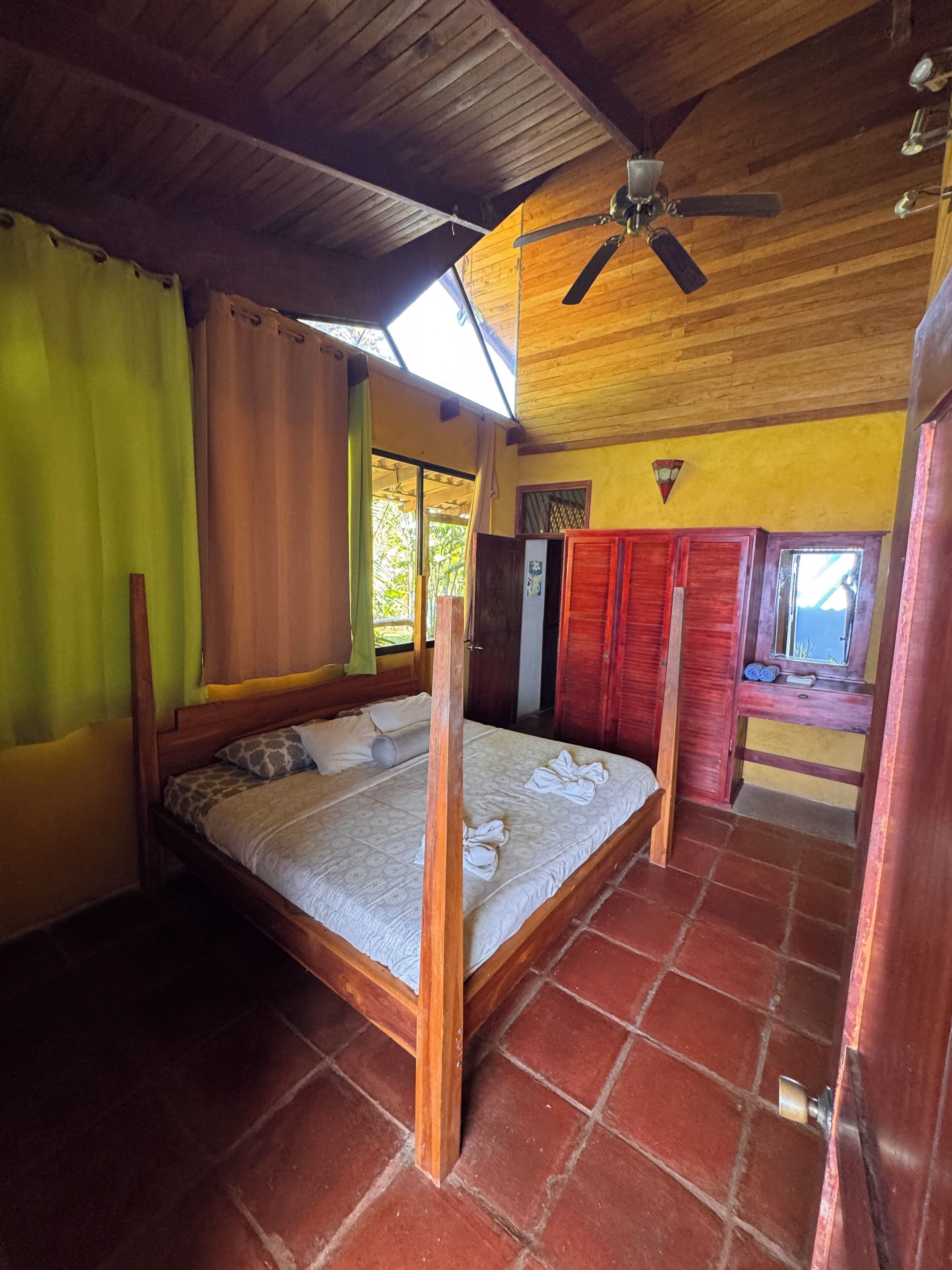 Um quarto com teto de madeira, cama com estrutura de madeira, ventoinha no teto e janela com cortinas.