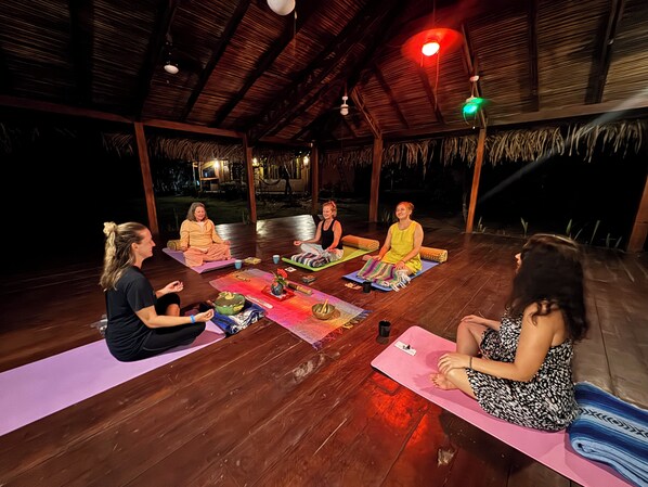 Un grupo de personas sentadas sobre esterillas de yoga en una habitación de madera con techo de paja.
