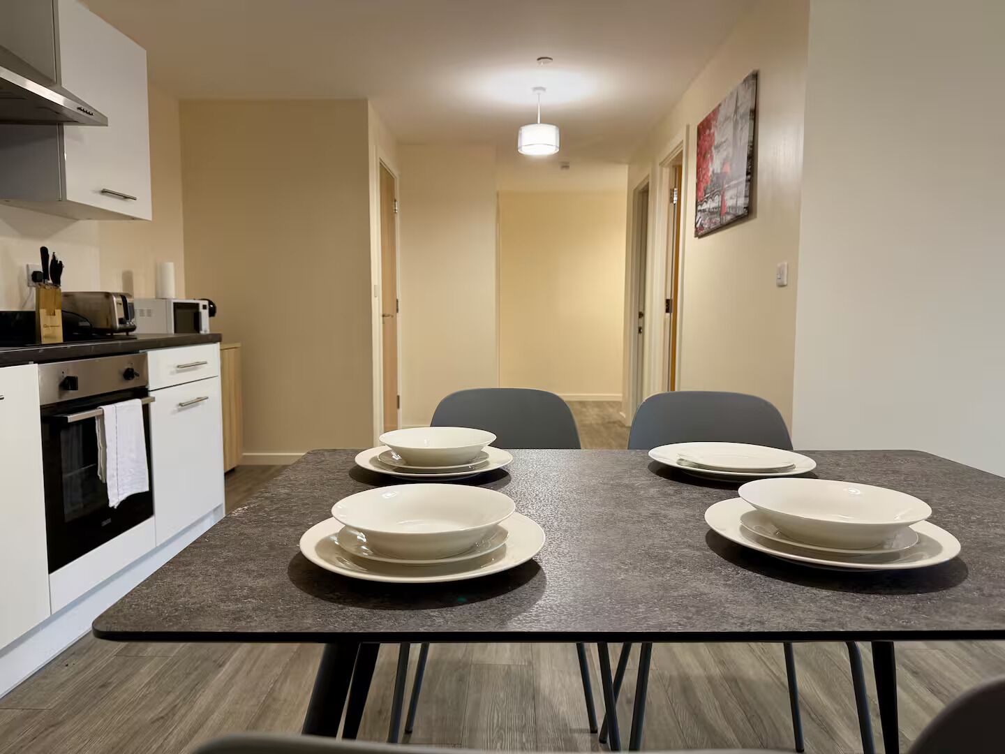 A modern kitchen with white cabinets, a built-in oven, and a microwave. There is a dining table with four white plates and two chairs.