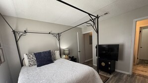 A four-poster bed with a white quilt, a dark throw pillow, and a bedside table with a lamp. A flat-screen TV is mounted on the wall to the right.