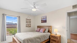 Ein Schlafzimmer mit Bett, Nachttisch mit Lampe, Deckenventilator, Fenster mit Vorhängen und einem Wandregal.