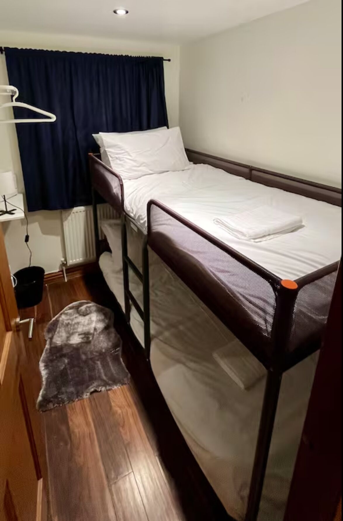 A single-bed room with a wooden floor, a small rug, a radiator, a window with dark curtains, and a white bedside table.