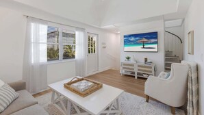 A spacious living room with a white sofa, a white coffee table, and a white armchair. There is a large window with white curtains, a painting of a beach scene, and a staircase leading to an upper level. - Beach Bungalow- 2 min walk to beach! Heated Pool (Bradenton Beach)
