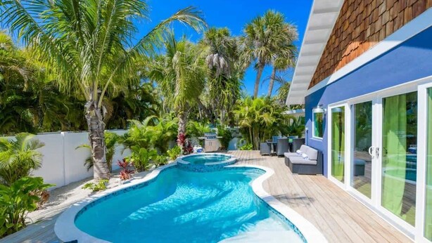 Ein Swimmingpool mit Holzterrasse, umgeben von Palmen und einem blauen Gebäude mit Glasüren.
