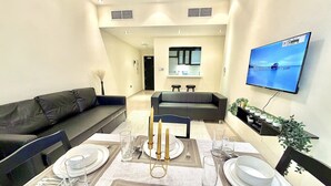 Un comedor con una mesa dispuesta para cuatro personas, un sofá de cuero negro, un televisor montado en la pared y una cocina al fondo.