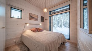 A cozy bedroom with a bed, two bedside lamps, a painting, and a large window with a snowy forest view. - Pitkäkuusen mökki, new cabin close to center of Äkäslompolo. Nature in backyard (Lappi)