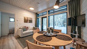 A cozy living room with a wooden interior, a dining table with a coffee table, and a large window offering a view of a snowy forest. - Pitkäkuusen mökki, new cabin close to center of Äkäslompolo. Nature in backyard (Lappi)