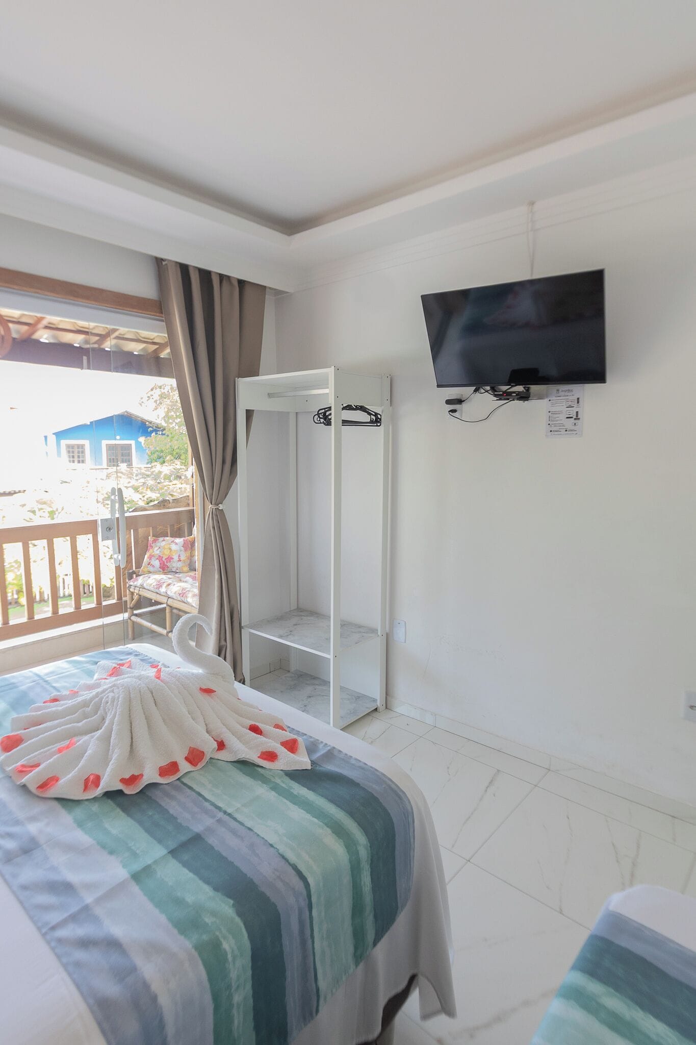 Quarto de hotel com cama, televisão presa na parede e janela com cortinas.