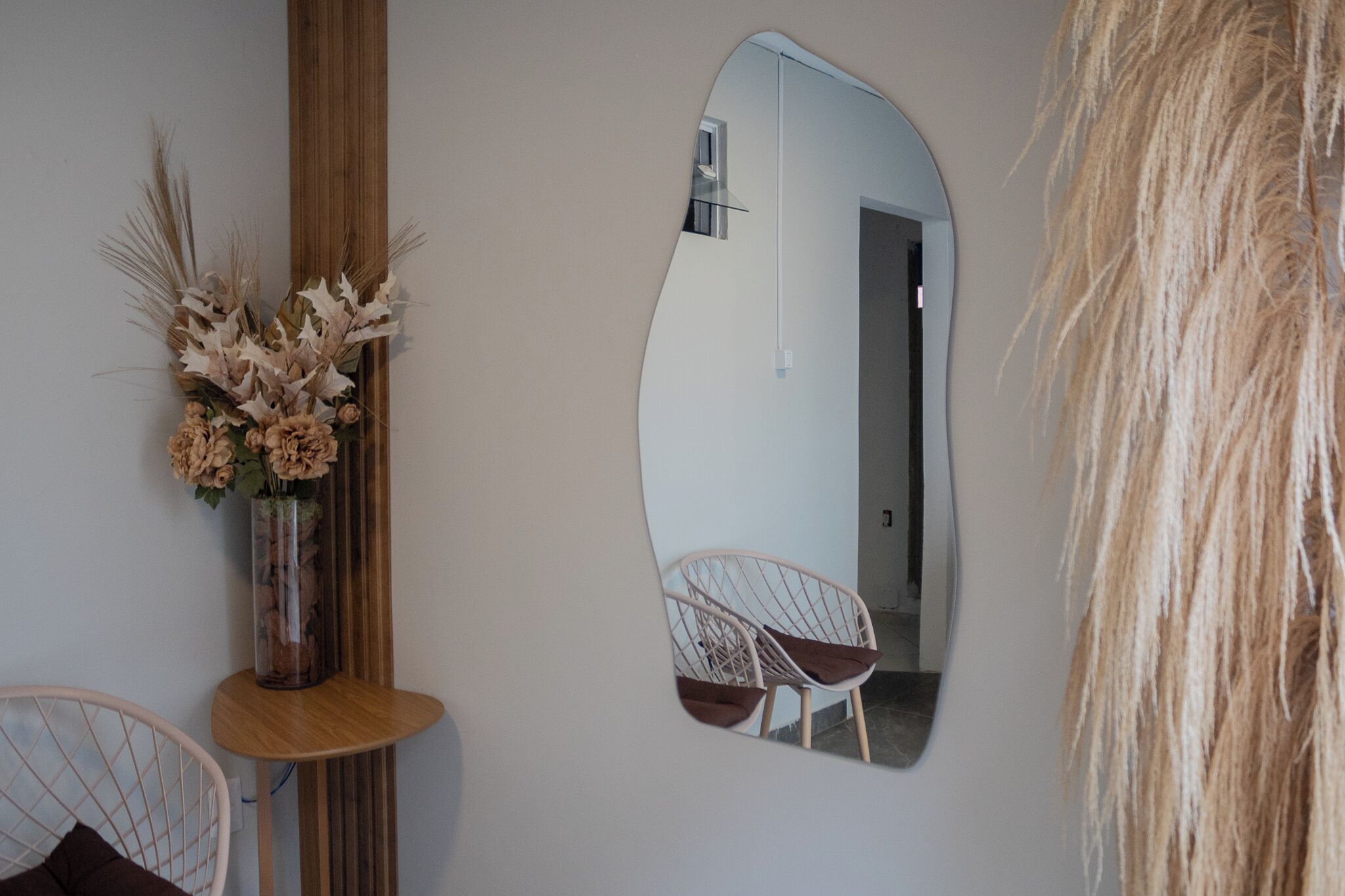 A room with a mirror reflecting a chair and a door, a vase with dried flowers, and a wooden side table.