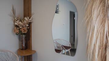 A room with a mirror reflecting a chair and a door, a vase with dried flowers, and a wooden side table.