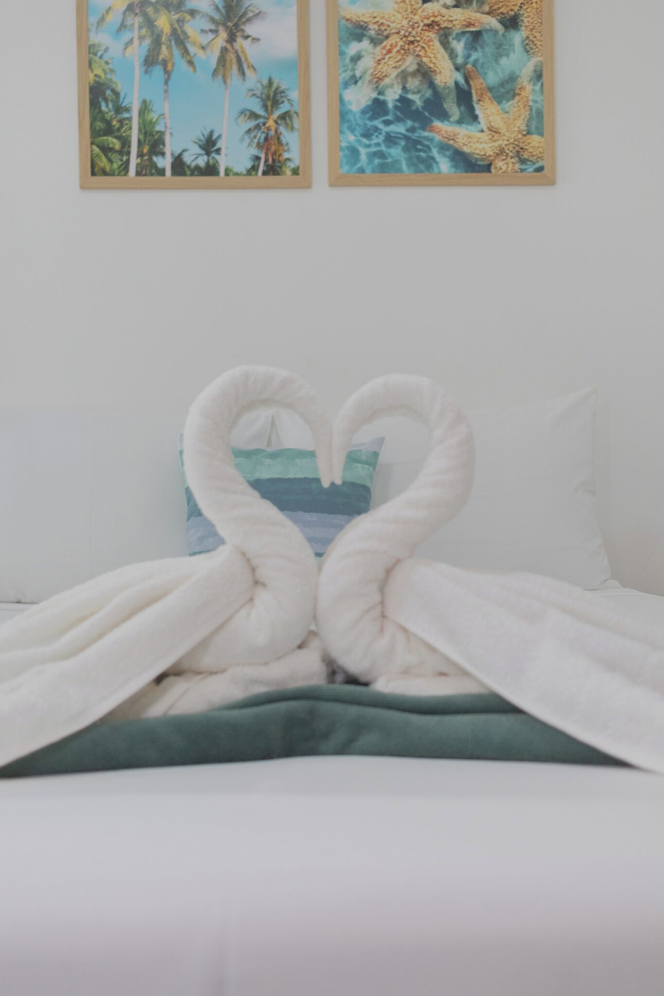 Un lit agrémenté d’une décoration en forme de cœur réalisée avec des serviettes, et un oreiller orné d’un motif représentant un paysage marin aux couleurs bleu et vert.