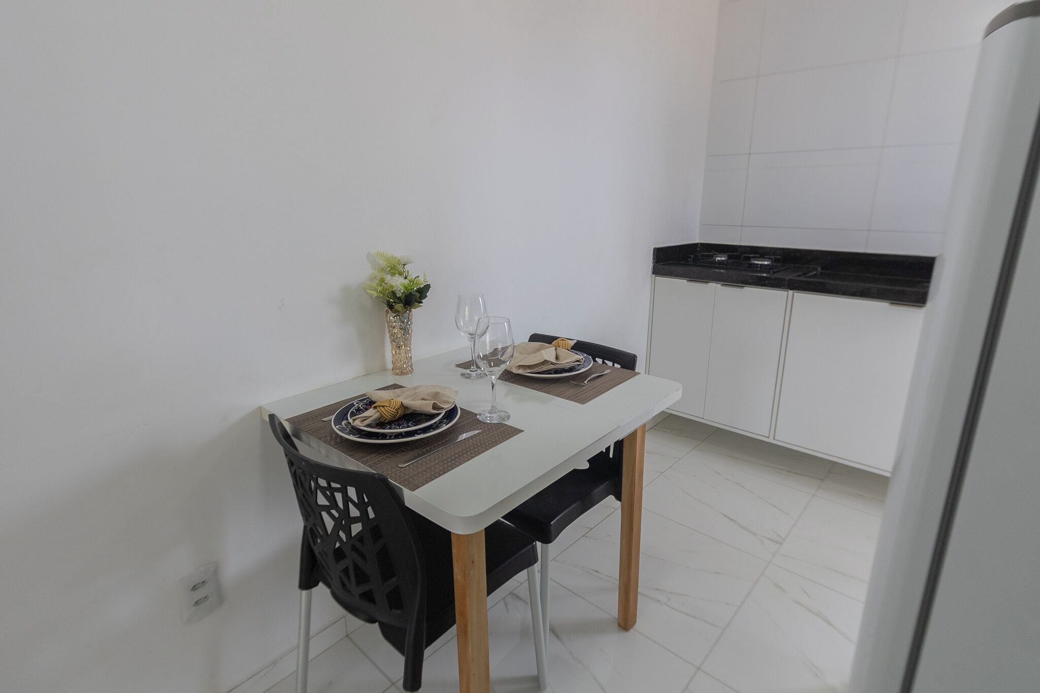Um espaço de refeições pequeno com uma mesa branca, duas cadeiras e um vaso com plantas verdes.