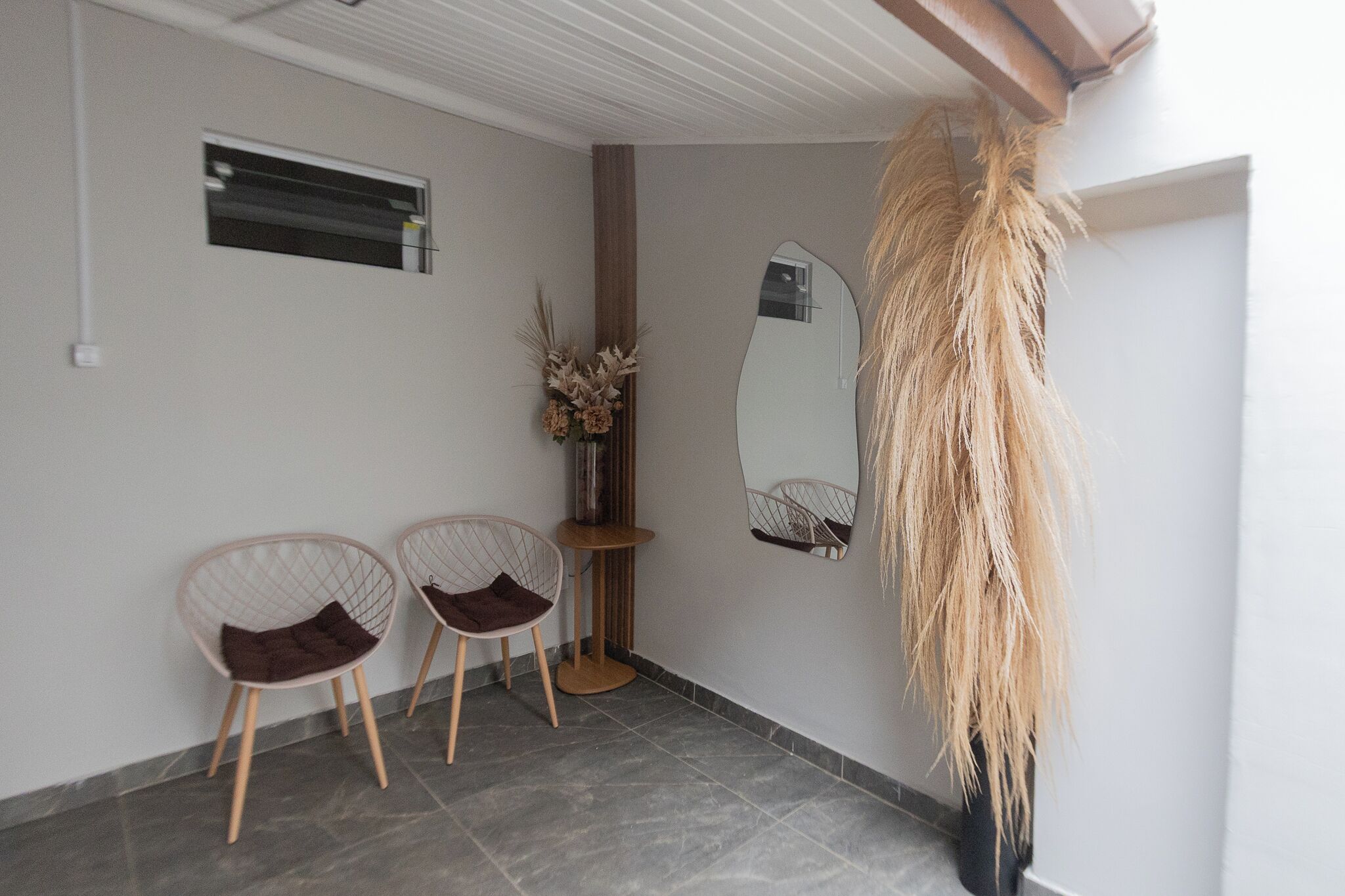 A room with two wireframe chairs, a small table with a vase of dried flowers, and a large mirror on the wall.