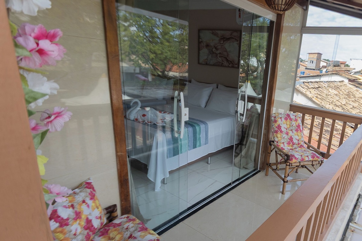 Ein Balkon mit einem Bett, einem Stuhl und einer Glastür, die zu einem Zimmer mit einem Bett und einem Gemälde führt.
