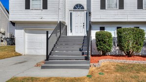 A two-story house with a front garage, a staircase with a black railing, and a small front yard with two bushes.