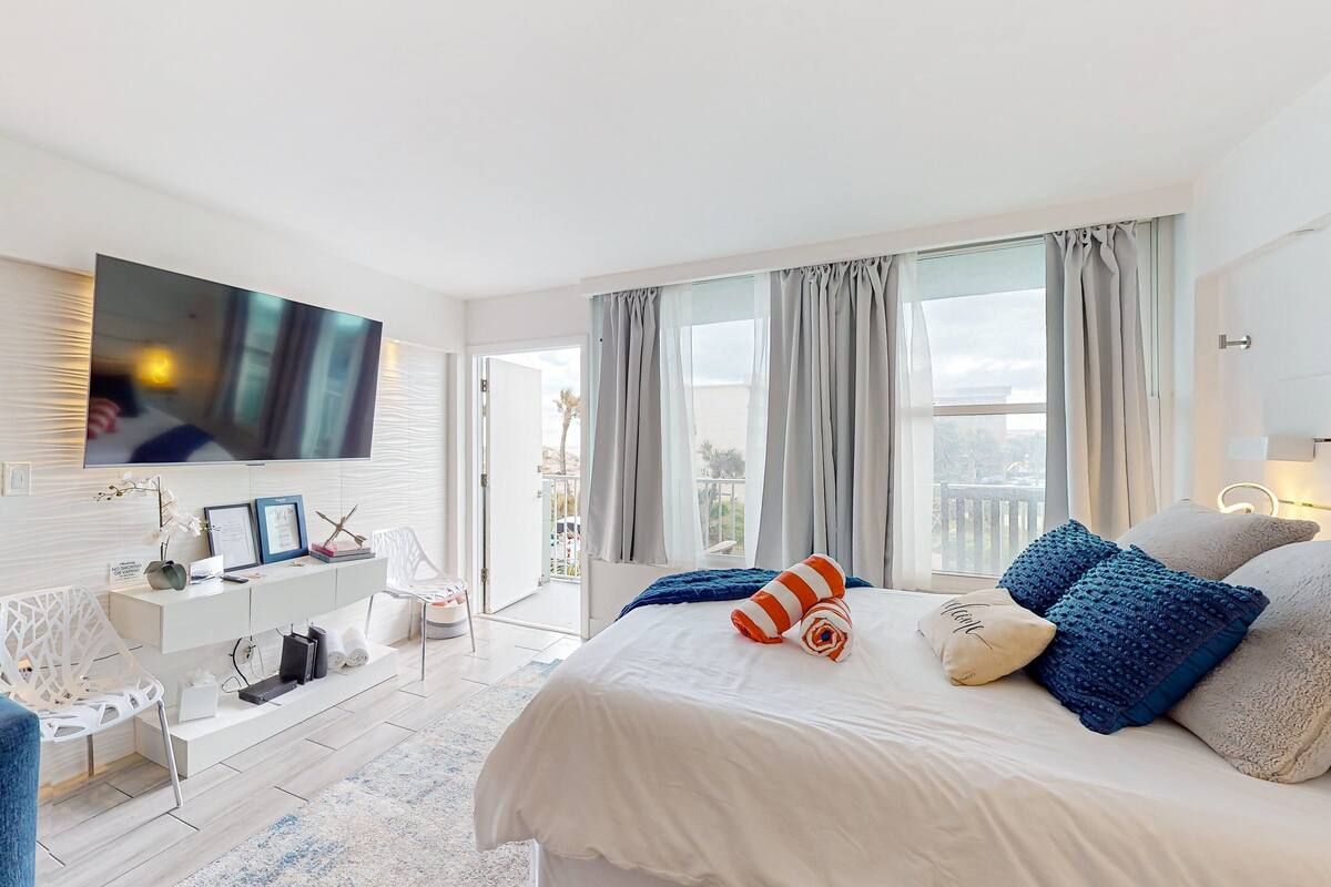 Ein modernes Schlafzimmer mit einem großen Bett, einem an der Wand montierten Flachbildfernseher, einem weißen Konsolentisch und einem Balkon mit Vorhängen.