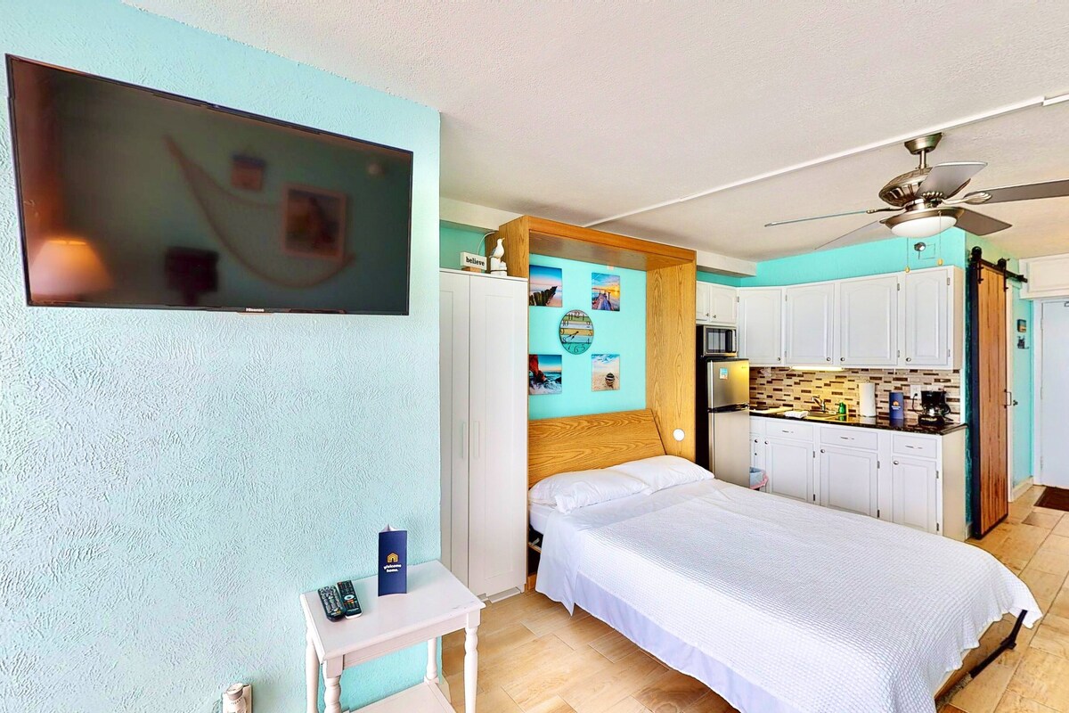 A bedroom with a bed, a small table, a TV, and a kitchenette.