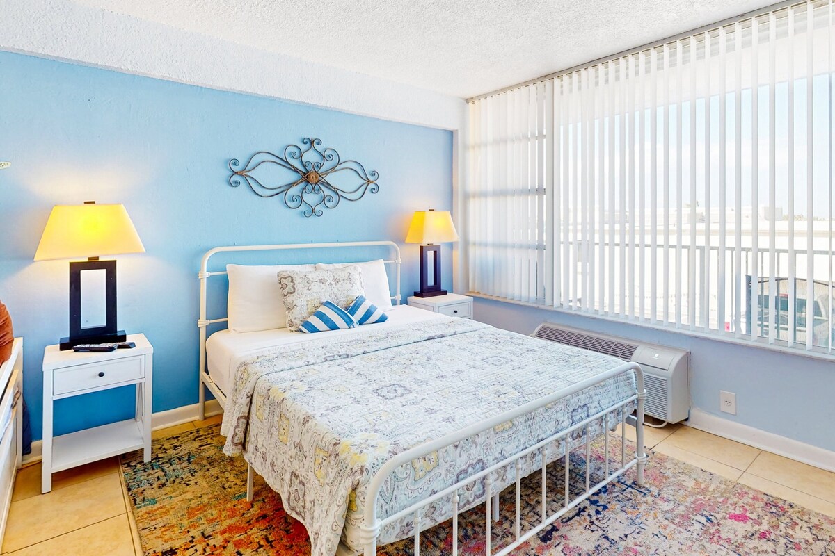 Un dormitorio con cama, dos mesitas de noche con lámparas, un adorno en la pared, un radiador y una ventana con persianas.