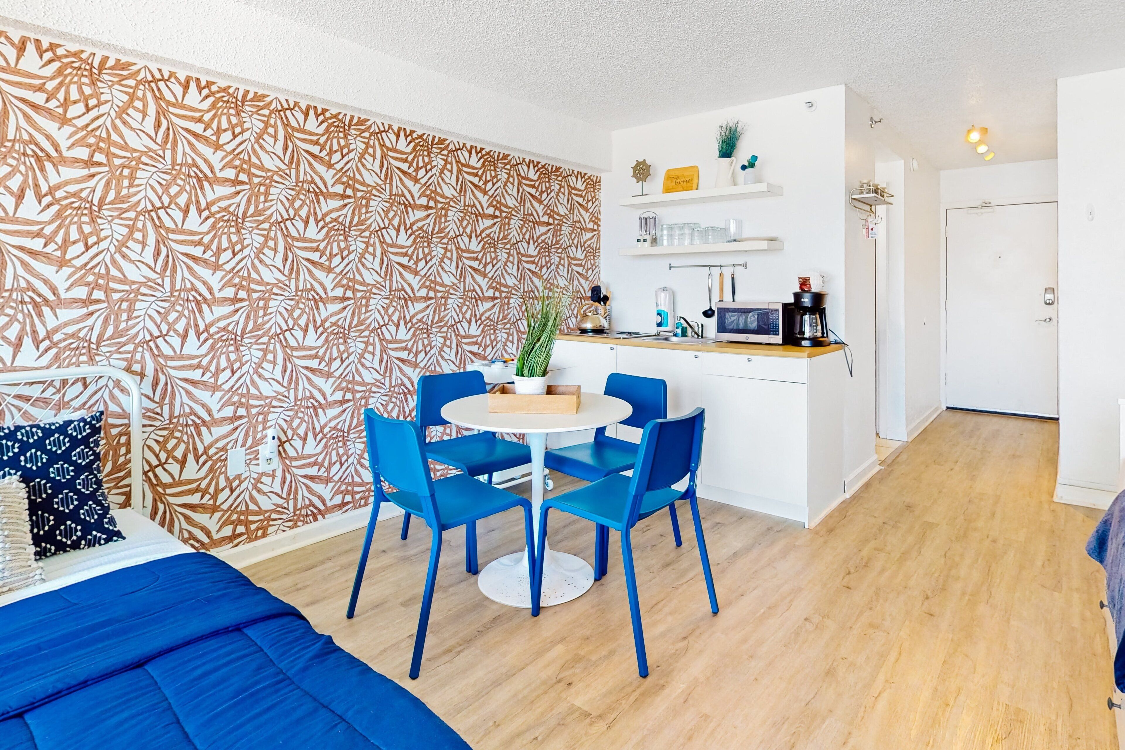 A compact living space with a bed, a dining area with blue chairs, a kitchen with a microwave, and a patterned wall.