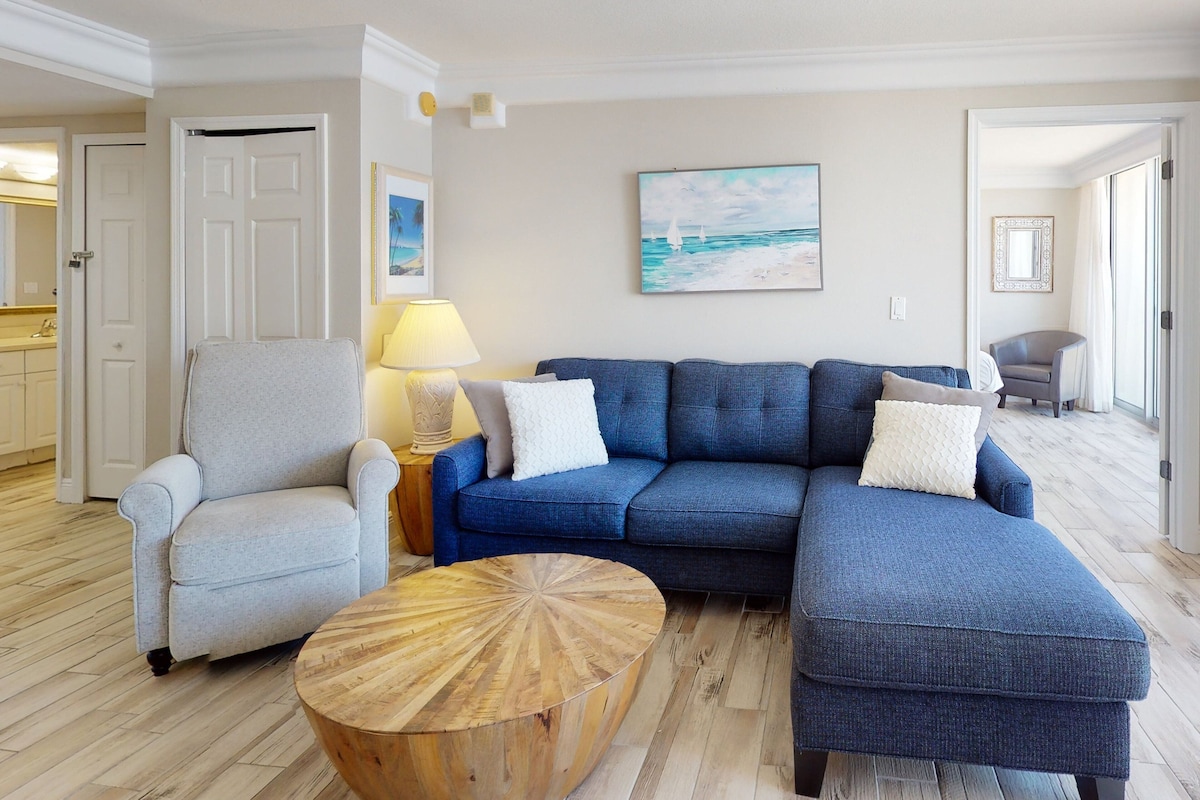 Una sala de estar con un sofá modular azul, un sillón gris, una mesa de centro de madera y un cuadro de veleros en la pared.