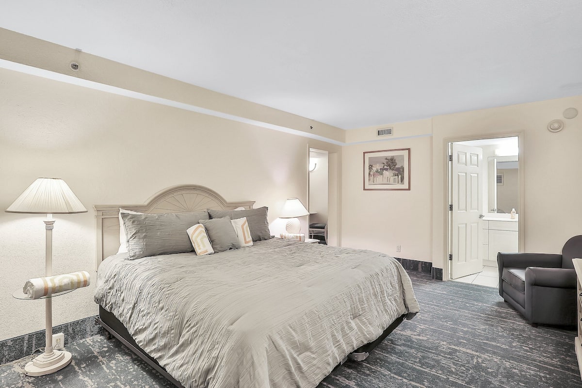 A hotel room with a large bed, two bedside lamps, a chair, and a door leading to a bathroom.