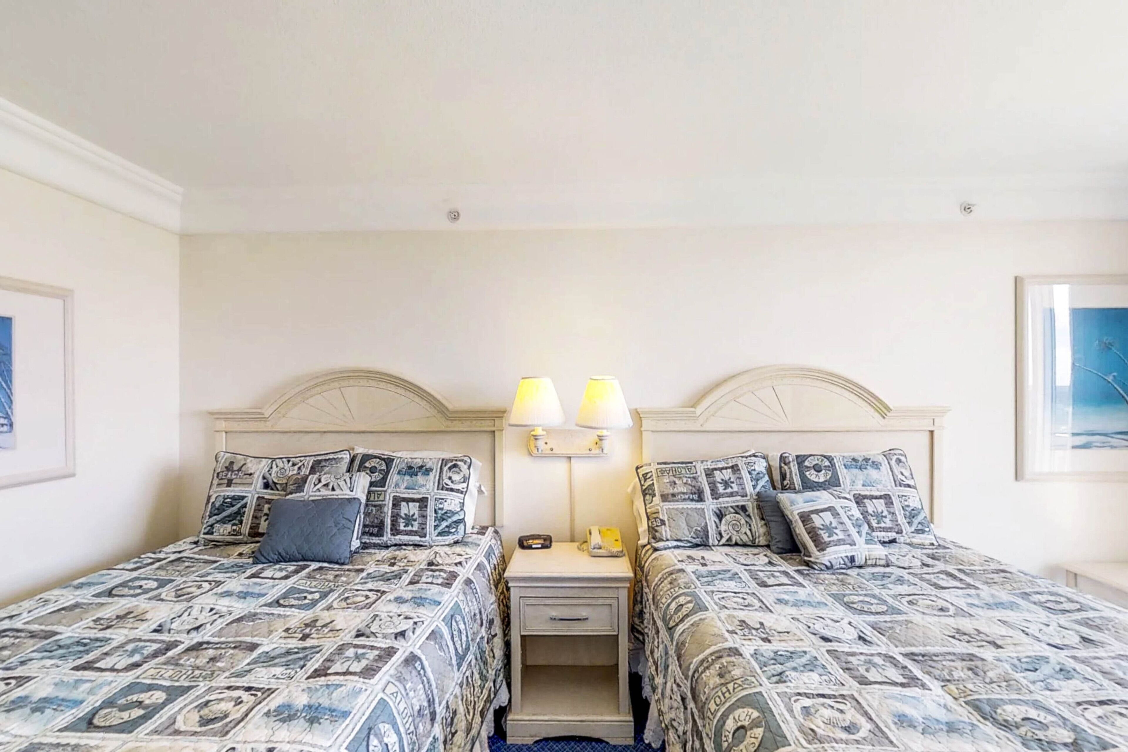 A hotel room with two beds, each with patterned bedding and matching pillows. There is a nightstand with a lamp between the beds.