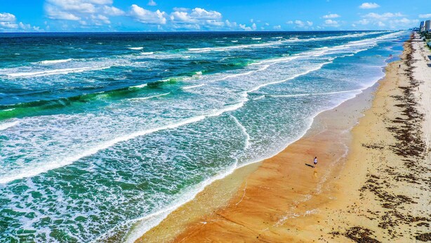 Ein Strand mit Wellen, eine Person, die spazieren geht, und Gebäude im Hintergrund.
