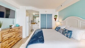 Ein Schlafzimmer mit einem Bett, einer Holzkommode, einem Fernseher und einem sichtbaren Badezimmer im Hintergrund.