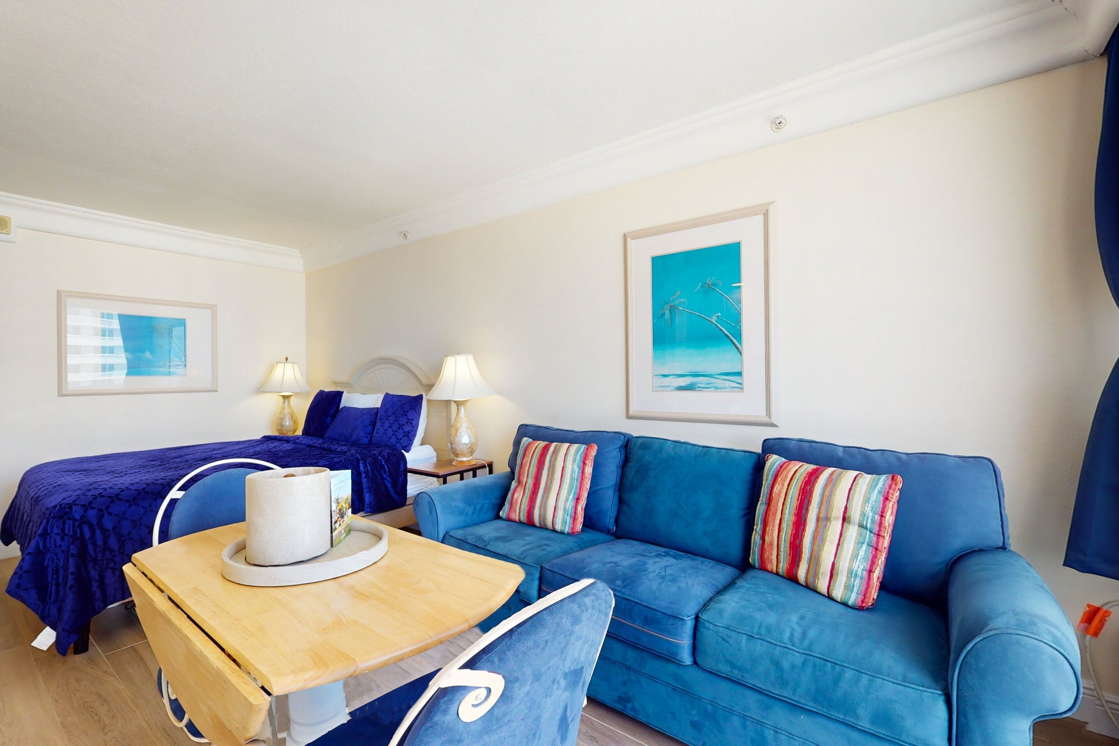 A room with a blue sofa, a bed with a blue cover, a wooden table, and two lamps.