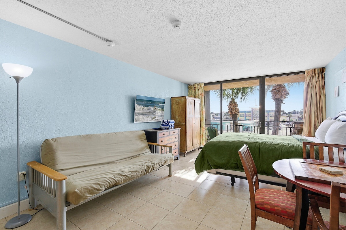 Habitación de hotel con cama, sofá, mesa de comedor y vistas a palmeras y al paisaje urbano.