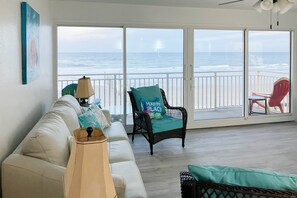 Ein Wohnzimmer mit einem weißen Sofa, einem Korbstuhl mit einem 'Happy Place'-Kissen, einer Lampe und Blick auf den Ozean durch große Schiebeglastüren.
