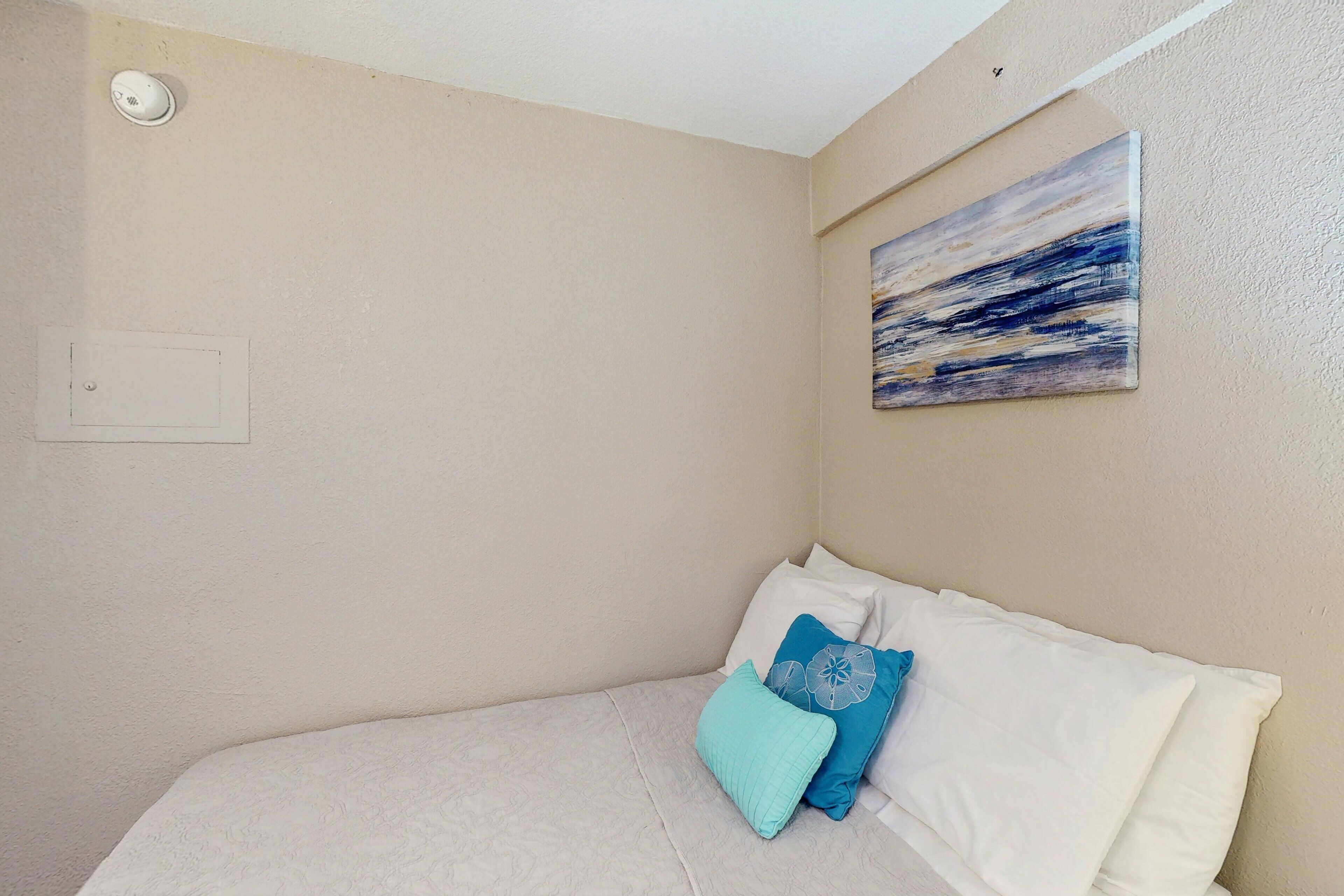 A bedroom with a bed, a painting, and a smoke detector.