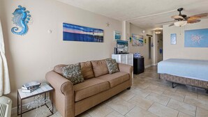 A living room with a beige sofa, a coffee table, a bed with a blue bedspread, and a ceiling fan. - Daytona Ocean Jewels (Daytona Beach)