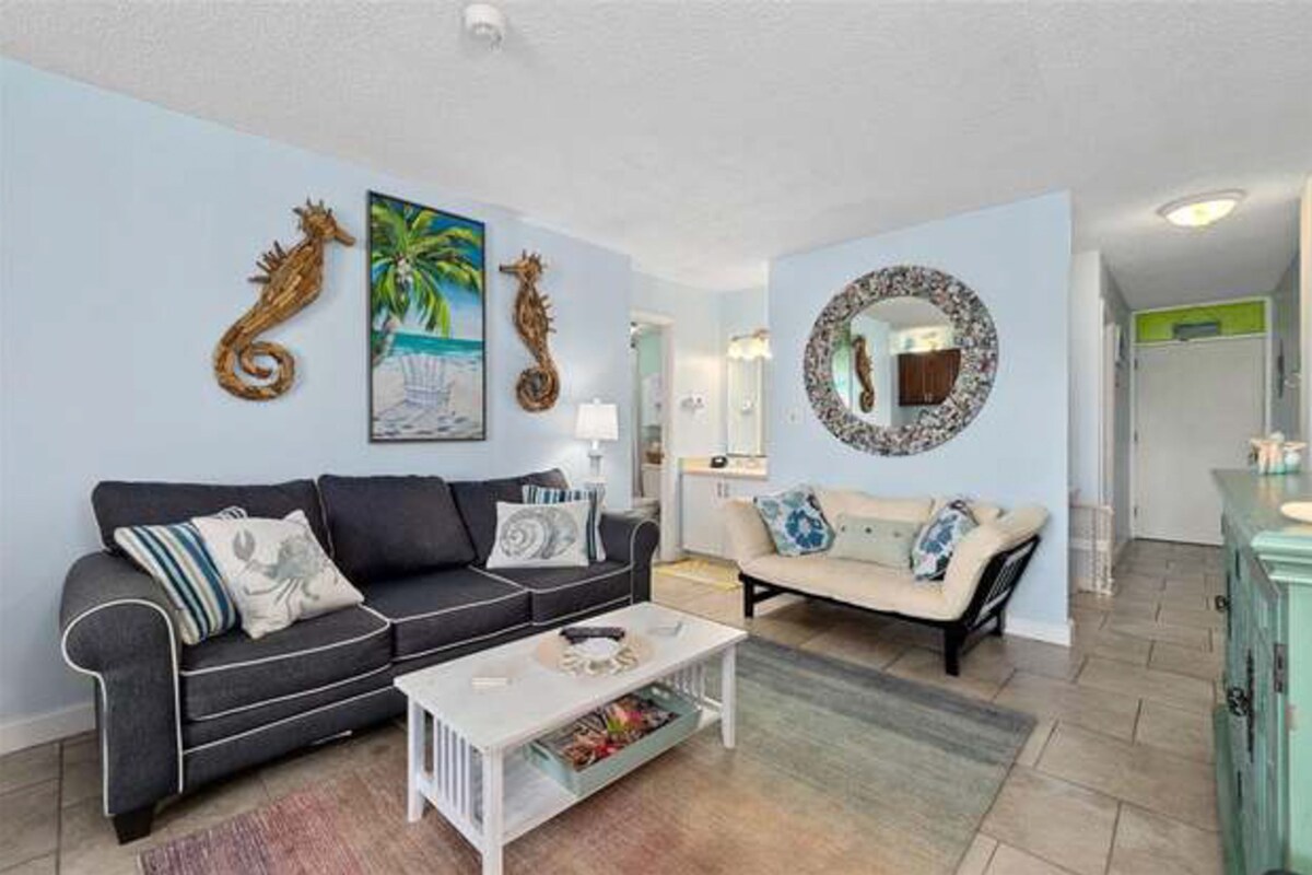 A living room with a dark grey sofa, a white coffee table, and a beige sofa. There are decorative seahorse wall art pieces and a round mirror with a seahorse design.