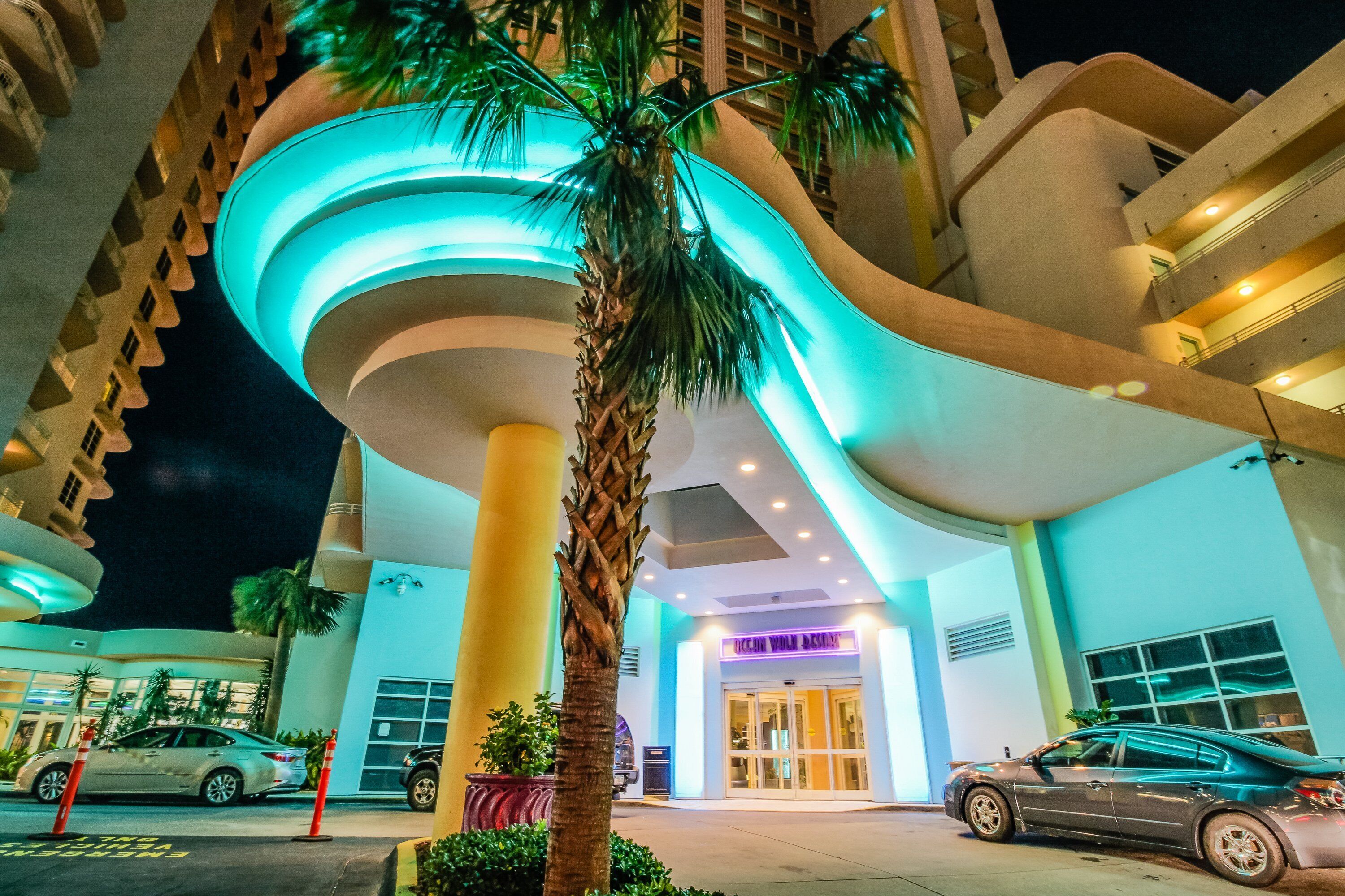 A modern hotel entrance with a curved facade, palm trees, and a parking area with cars.