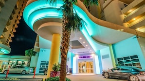 L’entrée d’un hôtel moderne, avec une façade incurvée, des palmiers et un espace de stationnement où sont garées des voitures.