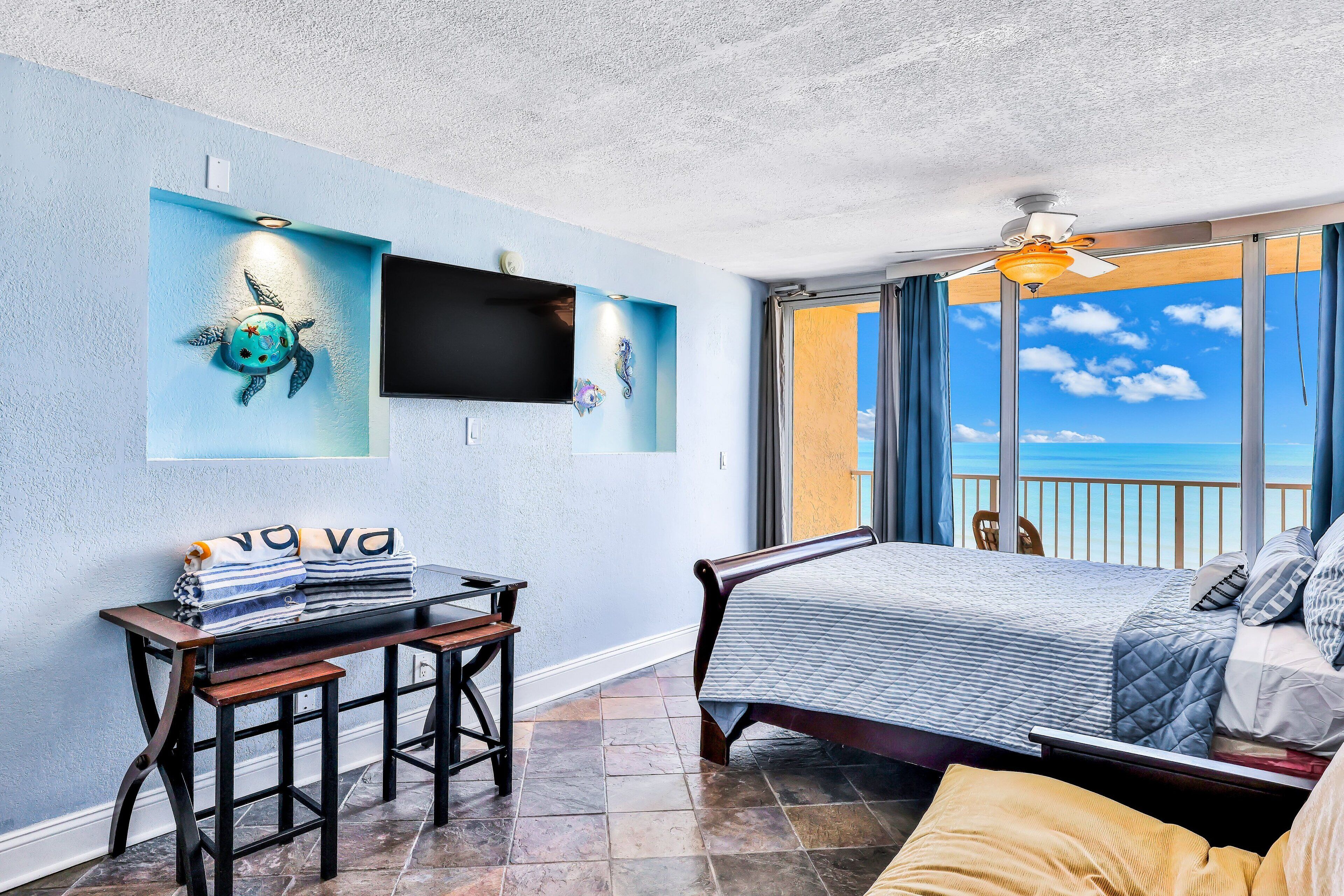 A bedroom with a bed, a desk, and a TV. There is a view of the ocean through large windows.