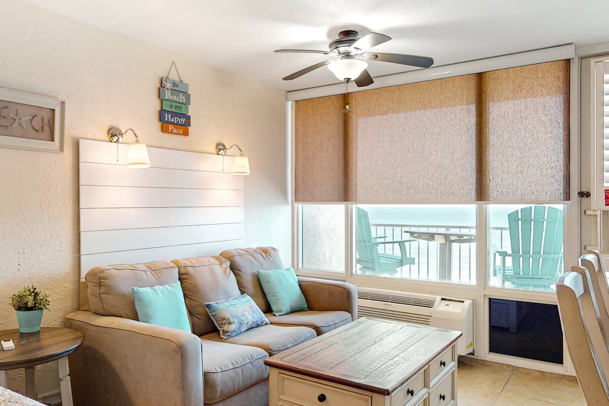 A cozy living room with a beige sofa, a wooden coffee table, and a ceiling fan. There is a large window with a view of outdoor furniture and a beach sign on the wall.