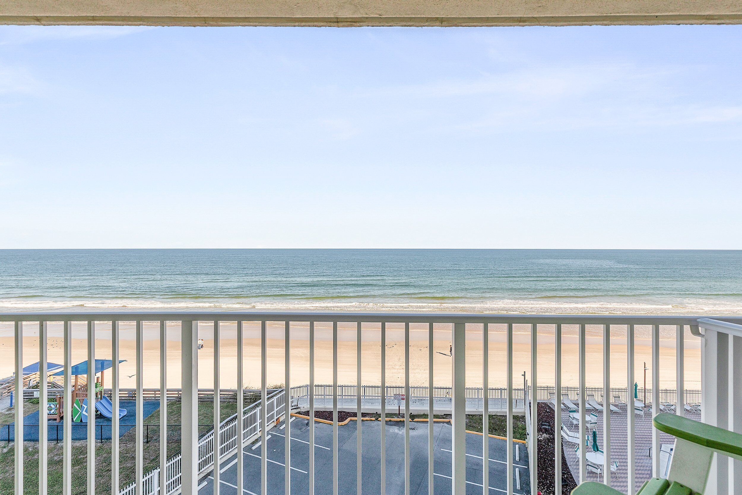 Une vue sur une plage, prise depuis un balcon doté d’une balustrade, avec une aire de jeux et un parking en arrière-plan.