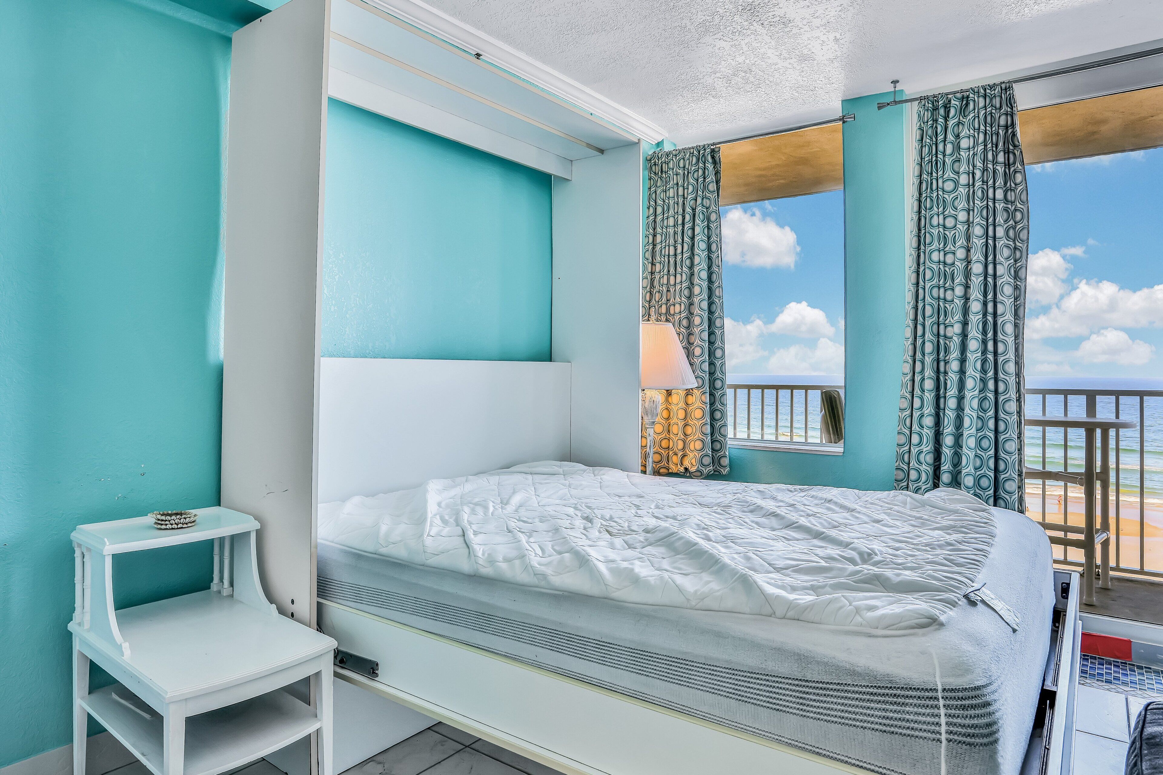 Ein Einzelbett mit weißer Matratze und Kopfteil, ein kleiner Nachttisch mit Lampe und Meerblick durch einen Balkon mit gemusterten Vorhängen.