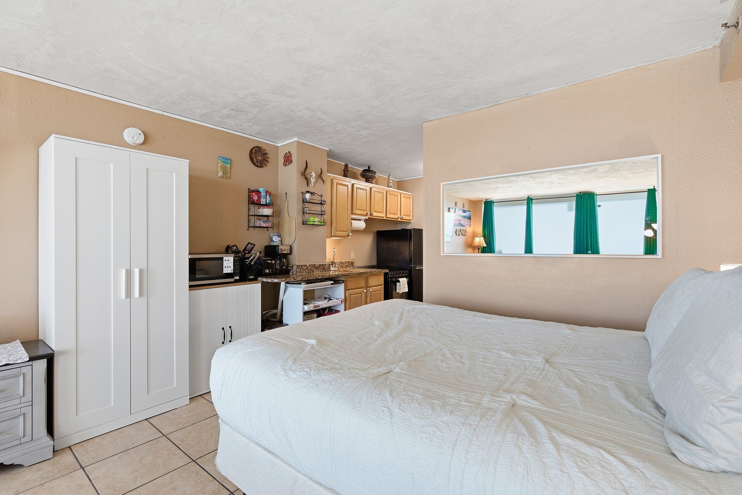 A bedroom with a large bed, a wardrobe, a kitchenette, and a window with curtains.