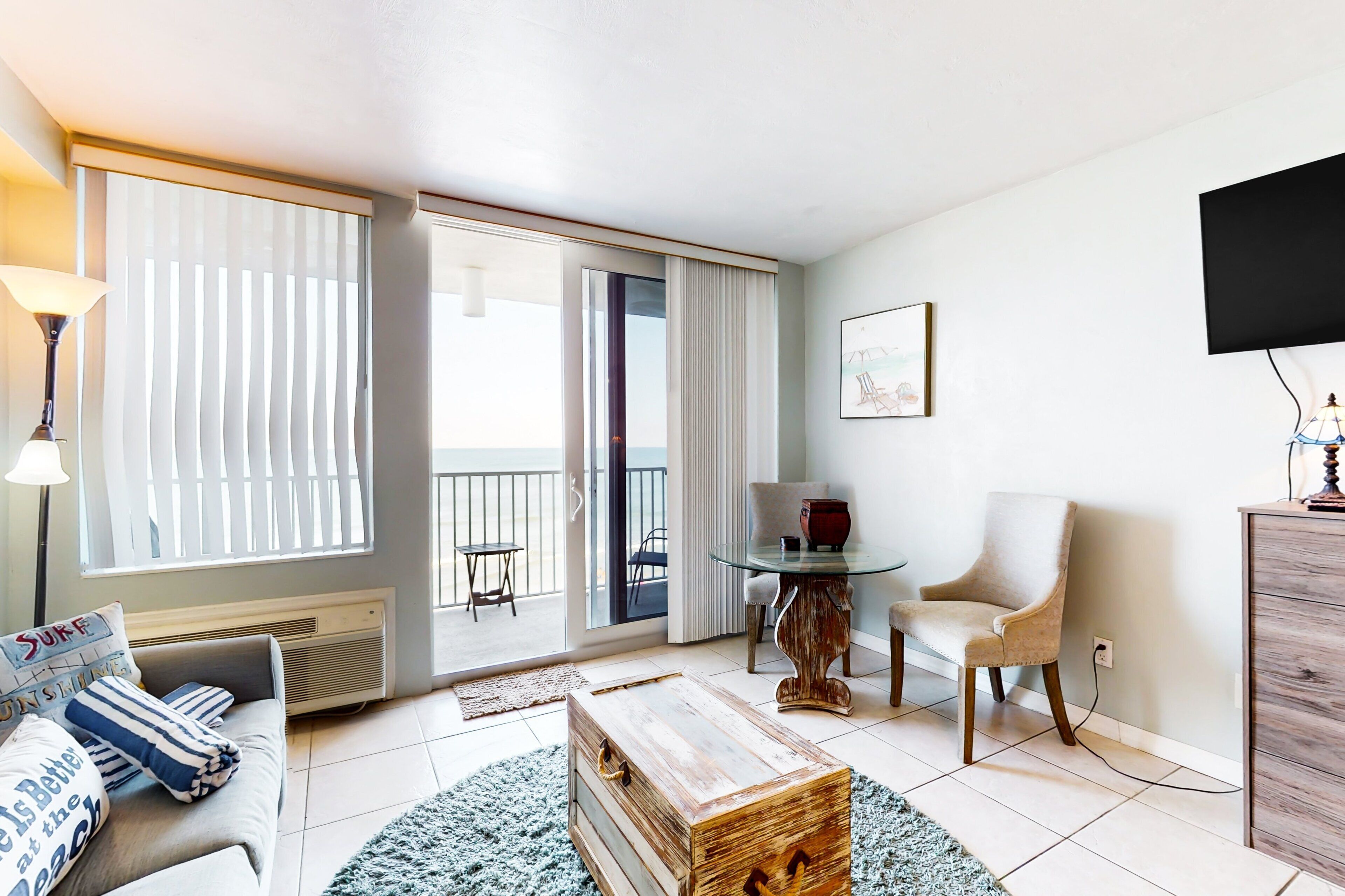 Ein modernes Wohnzimmer mit einer Couch, einem Holztisch, einem Glastisch und einem Sessel. Ein Balkon mit Blick, ein an der Wand montierter Fernseher und ein Heizgerät unter der Couch sind ebenfalls vorhanden.
