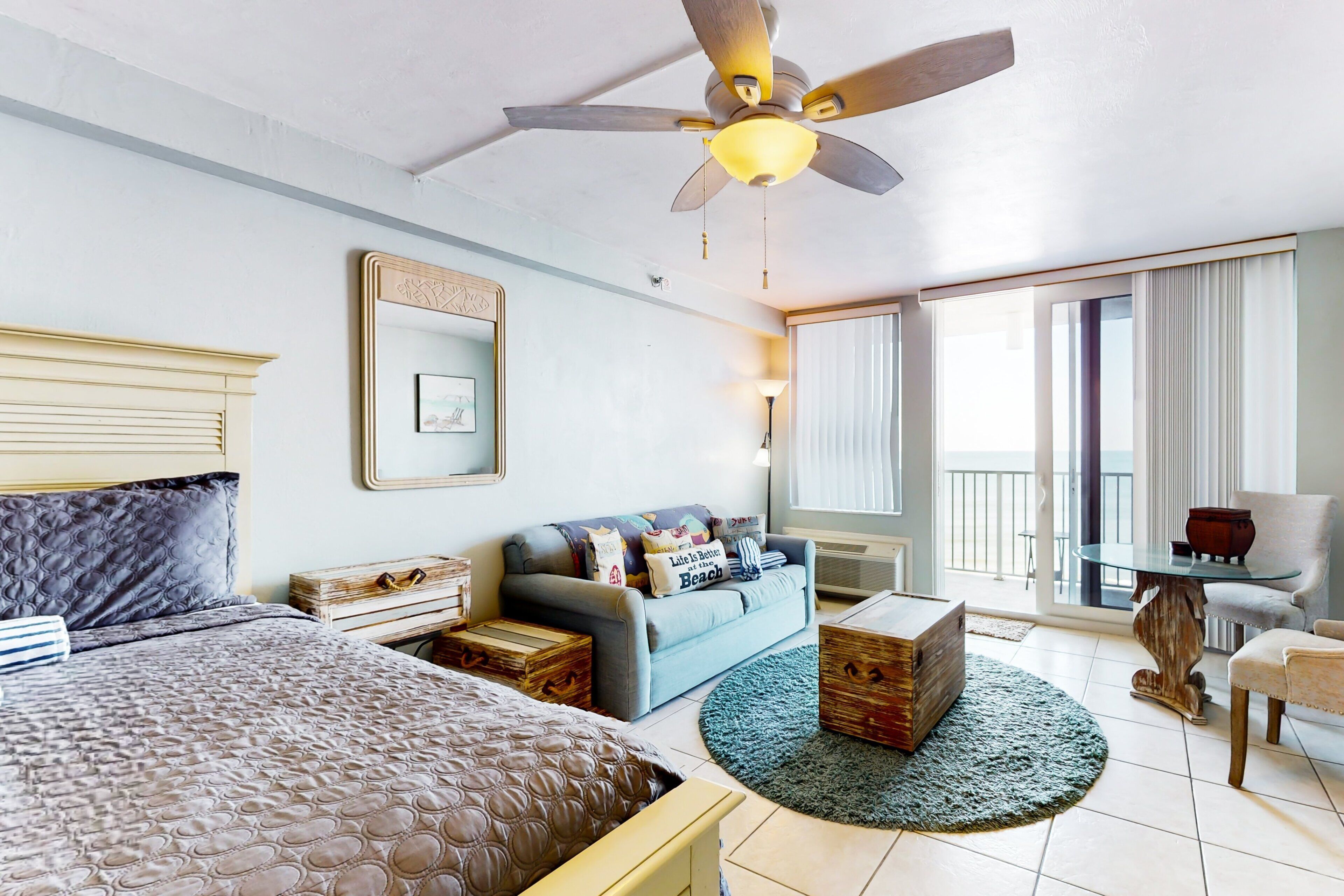 Habitación de hotel con cama, sofá, mesa de centro, sillón, ventilador de techo y ventana con cortinas.