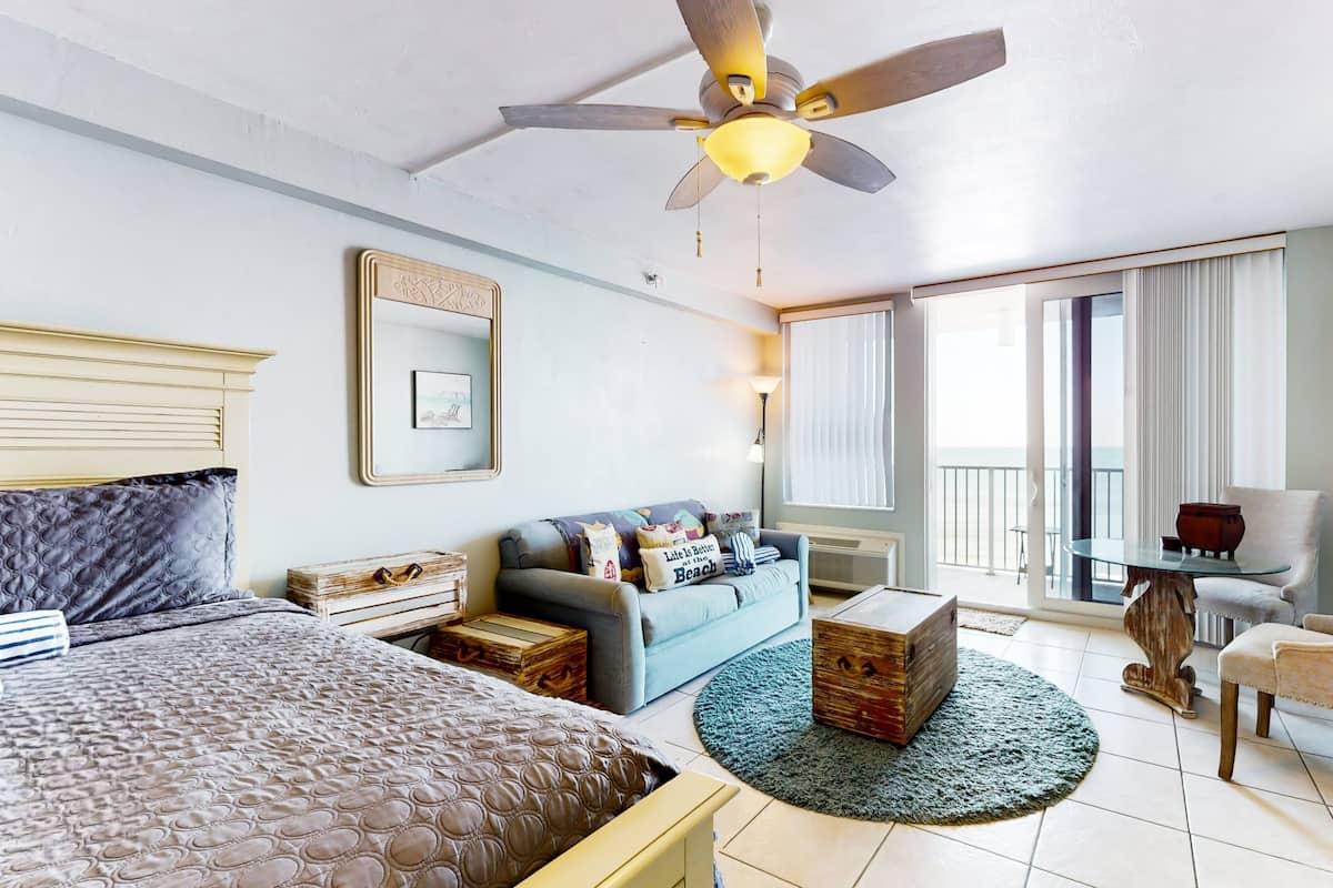 Habitación de hotel con cama, sofá, mesa de centro, sillón, ventilador de techo y ventana con cortinas.